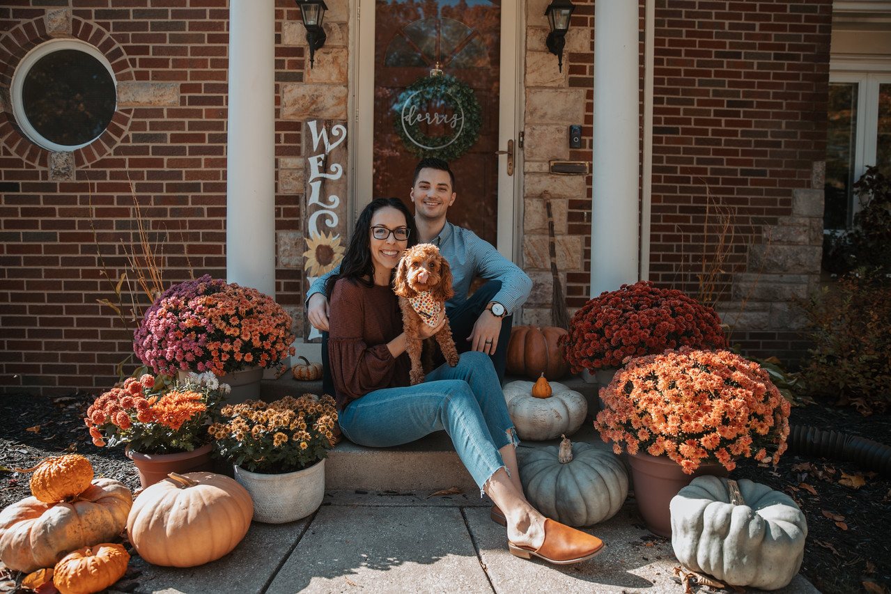 Alec Derrig with his wife and their dog on the front steps of their home, surrounded by fall mums and pumpkins